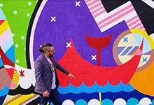 Man (jordan Bennett) standing in front of a colourful mural on a wall