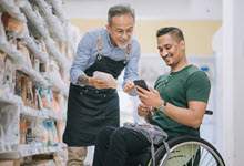 A person wearing a green t-shirt is sitting in a wheelchair at the grocery store, looking at his a phone smiling. Another person in a blue shirt is standing near, holding a package and smiling.