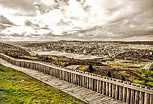 The photo shows green grass, a wooden walkway and a fence, in the background you can see the harbour and the city from a distance.