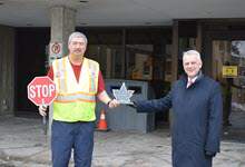 Crossing Guard holding a stop sign & wearing a safety vest is handed a glass trophy shaped like a maple leaf by the Mayor who is wearing a dark blue trenchcoat