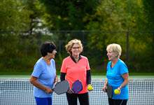 Three older women laughing & smiling stand in a pickleball (tennis) court holding pickleball paddles and ball.