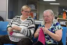 two women sitting near each other while knitting, one woman glances her friends knitting project
